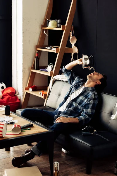 Hombre beber agua de la tetera mientras está sentado en el sofá en la sala de estar desordenada - foto de stock