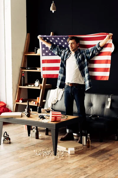 Bel homme tenant drapeau américain dans le salon désordonné — Photo de stock