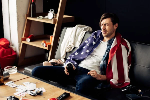 Uomo assonnato che tiene lattina con birra e seduto con bandiera americana sul divano — Foto stock
