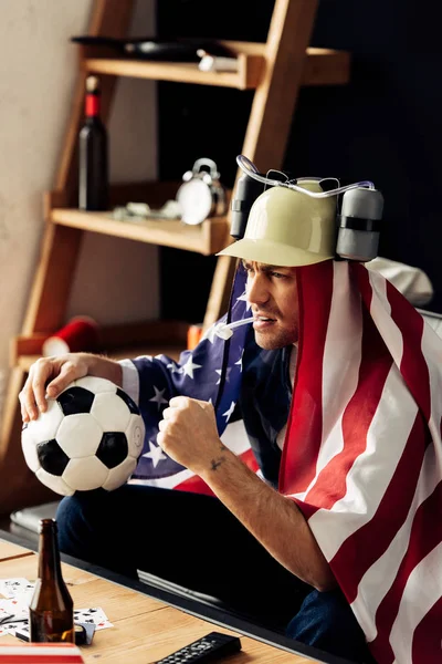 Homme portant un casque de bière regarder jeu avec drapeau américain sur les épaules — Photo de stock