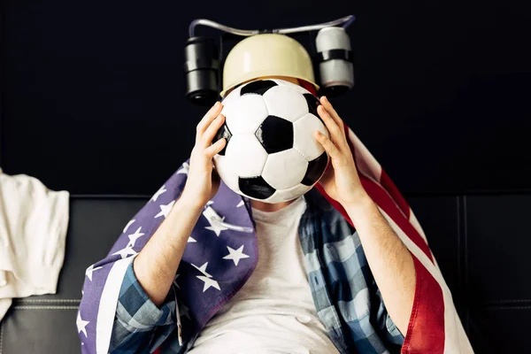Hombre con casco de cerveza cubriendo la cara con el fútbol y sentado con bandera americana en los hombros - foto de stock