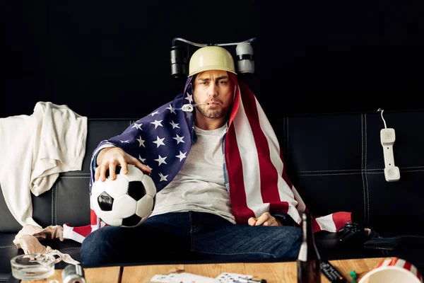 Uomo che indossa il casco di birra bere e tenere il calcio mentre seduto con bandiera americana sulle spalle — Foto stock