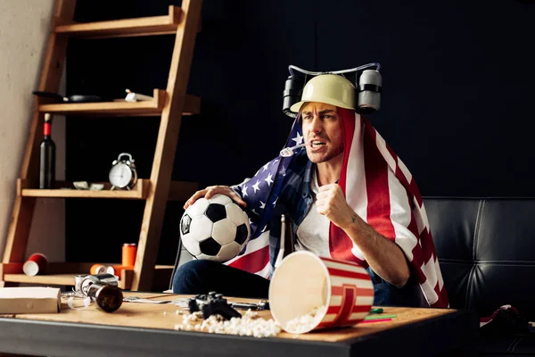 Man wearing beer helmet drinking and holding ball while sitting with american flag on shoulders and watching game — Stock Photo