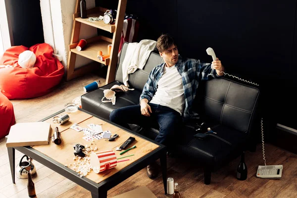 Пьяный мужчина смотрит на ретро-телефон и сидит на диване в грязном доме — стоковое фото