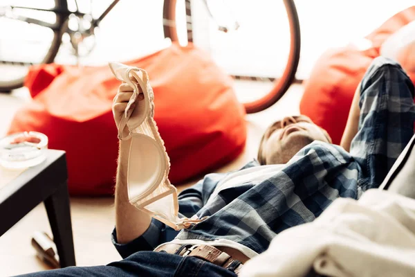 Foco seletivo do homem segurando sutiã enquanto deitado no sofá após a festa — Fotografia de Stock