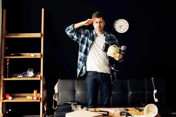 Hombre guapo sosteniendo casco de cerveza en las manos y de pie en la sala de estar desordenado - foto de stock