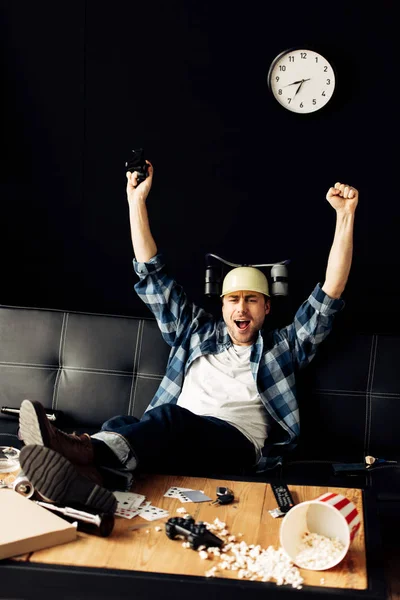 Hombre feliz con casco de cerveza celebrando la victoria en la sala de estar desordenada - foto de stock