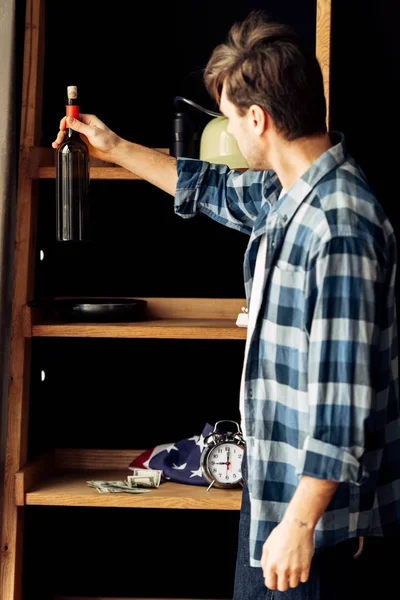 Uomo in possesso di bottiglia di vino vicino rack in soggiorno — Foto stock