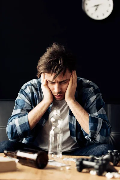 Man suffering from hangover holding head and looking at bottle at home — Stock Photo