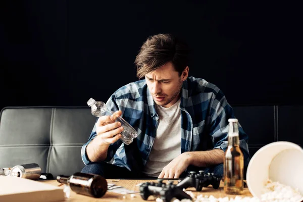 Hombre guapo que sufre de resaca mirando a la botella vacía mientras está sentado en el sofá - foto de stock