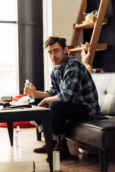 Schöner Mann schaut in die Kamera und hält Flasche, während er auf dem Sofa sitzt — Stockfoto