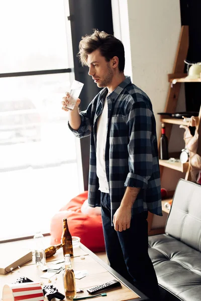 Уставший мужчина держит стакан воды возле кофейного столика с бутылками дома — стоковое фото