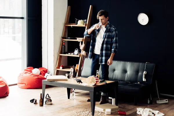 Drunk man holding glass and drinking water after party in messy living room — Stock Photo