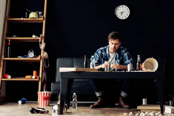 Hombre sosteniendo aspirina cerca de un vaso de agua en la sala de estar desordenada - foto de stock