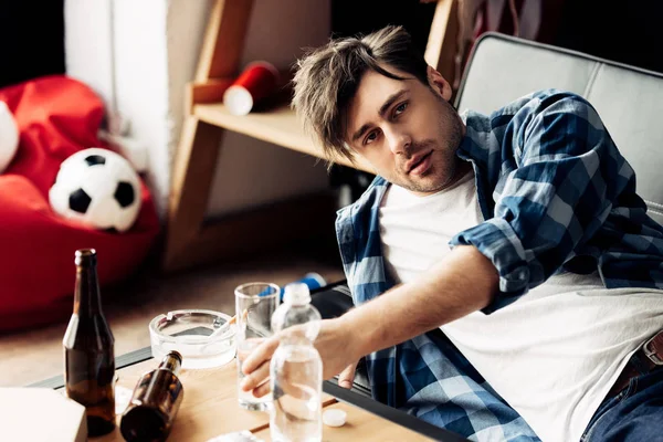 Foco seletivo do homem cansado tomando copo de água na mesa de café enquanto deitado no sofá — Fotografia de Stock