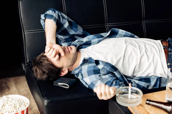 Hombre agotado acostado en el sofá y cubriendo la cara con la mano después de la fiesta en casa - foto de stock