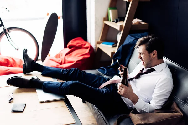 Mann im Anzug hält Flasche in der Hand und schreit nach Smartphone, während er nach Party auf Sofa sitzt — Stockfoto