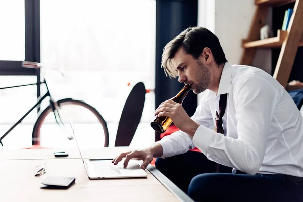 Tired man in suit drinking beer from bottle while using laptop after party — Stock Photo