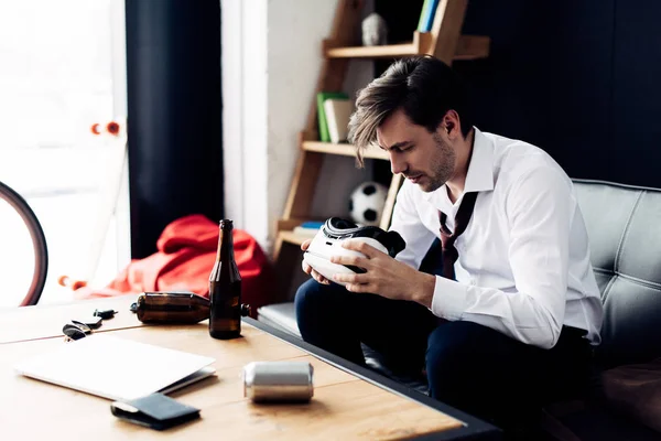 Hombre soñoliento mirando los auriculares de realidad virtual mientras está sentado en el sofá después de la fiesta - foto de stock
