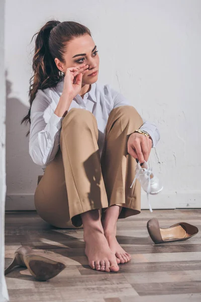 Расстроена женщина сидит на полу, плачет и держа детские туфли возле белой стены дома, скорбящее расстройство концепции — стоковое фото