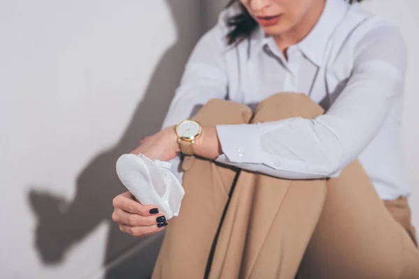 Vista recortada de la mujer triste en blusa blanca y pantalones beige sentado en el suelo cerca de la pared y la celebración de zapatos de bebé en casa, concepto de trastorno de duelo - foto de stock