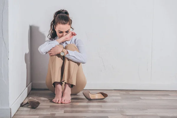 Femme triste en chemisier blanc et pantalon beige assis sur le sol et pleurant près du mur à la maison, concept de trouble de deuil — Photo de stock