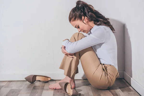 Mujer triste en blusa blanca y pantalones beige sentado en el suelo con la cabeza doblada cerca de la pared blanca en casa, concepto de trastorno de duelo - foto de stock