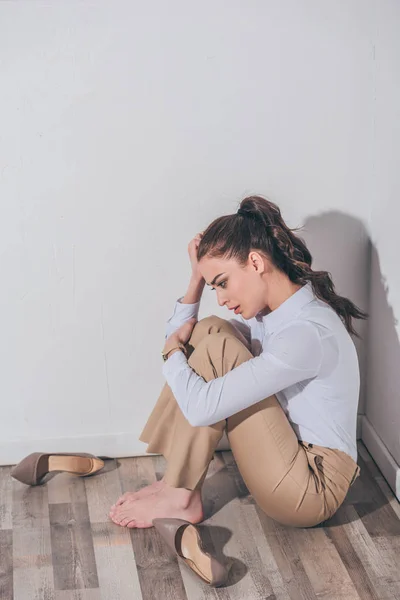 Traurige Frau in weißer Bluse und beiger Hose, die zu Hause auf dem Boden neben der Wand sitzt und trauert. — Stockfoto