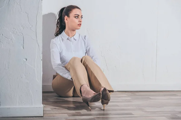 Засмучена жінка в білій блузці і бежеві штани, що сидять на підлозі біля білої стіни вдома, сумна концепція розладу — стокове фото