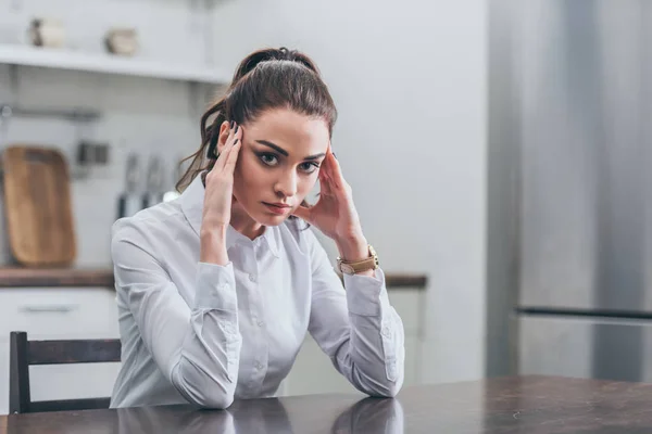 Upset woman in white blouse sitting at table and putting hands forehead in kitchen, grieving disorder concept — Stock Photo