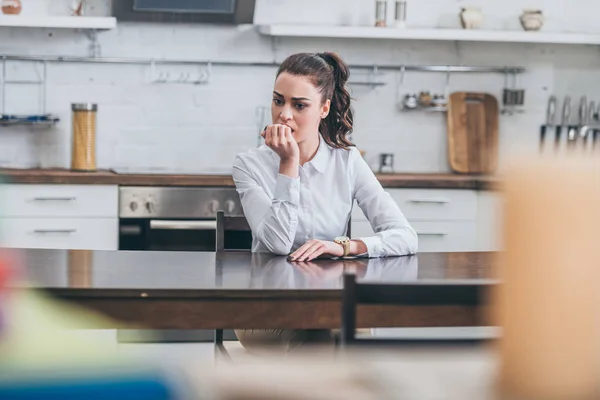 Upset woman in white blouse sitting ar table and bitting nails in kitchen, grieving disorder concept — Stock Photo