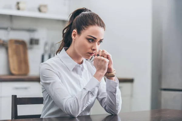 Upset woman in white blouse sitting at table in kitchen and thinking, grieving disorder concept — Stock Photo