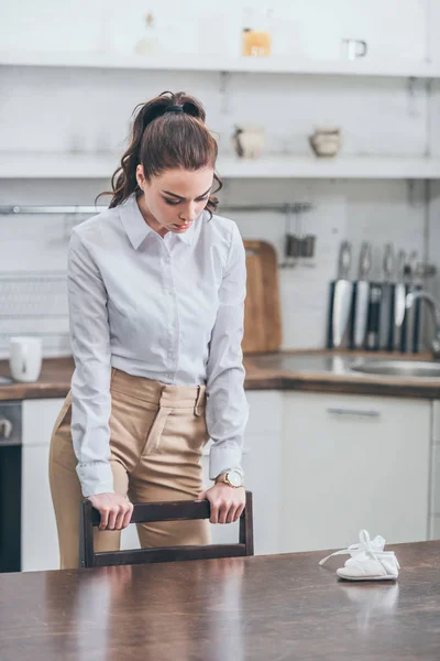 Засмучена жінка в білій блузці стоїть за столом на кухні, думаючи і дивлячись вниз, сумуючи за концепцією розладу — стокове фото