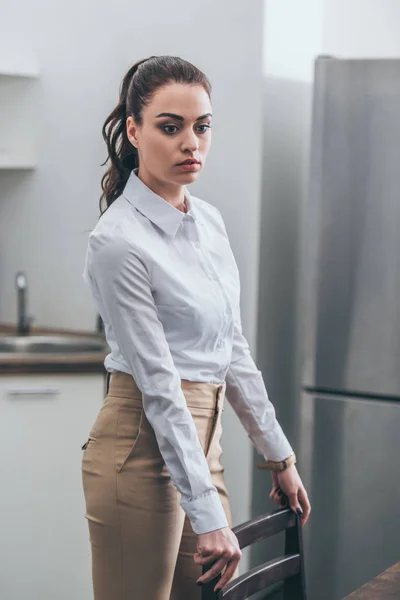 Upset woman in white blouse and beige pants standing by chair in kitchen, grieving disorder concept — Stock Photo