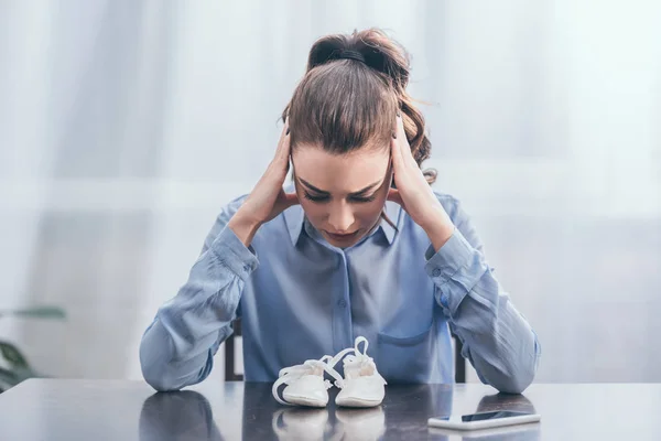 Sad woman in blue blouse sitting at wooden table with smartphone and looking at white baby shoes at home, grieving disorder concept — Stock Photo