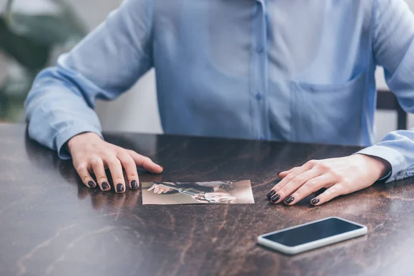 Обрізаний вид на жінку в блакитній блузці, сидячи за дерев'яним столом з смартфоном і фото вдома, сувора концепція розладу — стокове фото