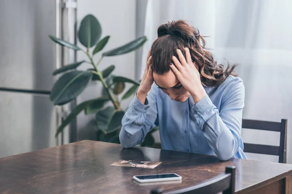 Mujer triste en blusa azul sentada en la mesa de madera con teléfono inteligente y mirando la foto en la habitación, concepto de trastorno de duelo - foto de stock