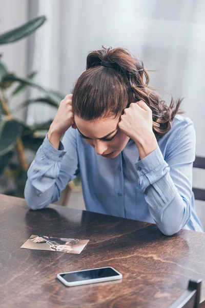 Mulher triste em blusa azul sentado à mesa com smartphone e olhando para a foto em casa, conceito de transtorno de luto — Fotografia de Stock
