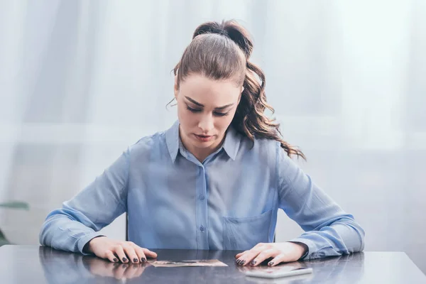 Mujer molesta en blusa azul sentada en la mesa de madera con teléfono inteligente y mirando la foto en la habitación, concepto de trastorno de duelo - foto de stock