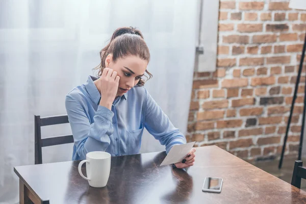 Upset woman in blue blouse sitting at table with smartphone, white cup and looking at photo at home, grieving disorder concept — Stock Photo