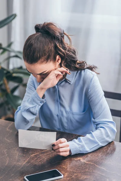 Aufgebrachte Frau in blauer Bluse sitzt mit Foto am Tisch, Smartphone und weint zu Hause, Konzept der Trauerstörung — Stockfoto