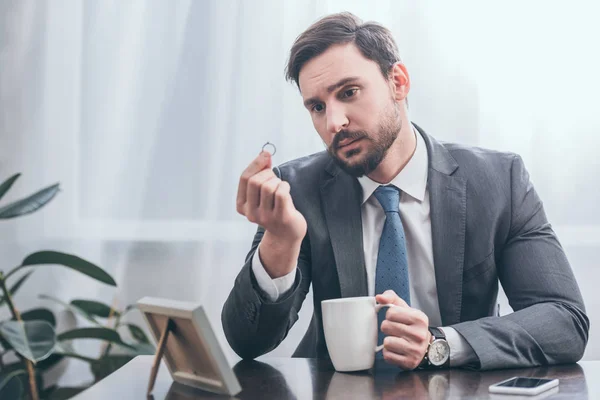 Sad man in gray suit sitting at wooden table with smartphone, photo in frame, holding white cap and looking at ring in room, grieving disorder concept — Stock Photo