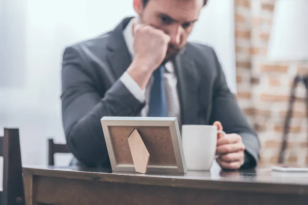 Enfoque selectivo del hombre molesto en traje gris sentado en la mesa de madera, sosteniendo la taza blanca y mirando el marco de la foto en la habitación, concepto de trastorno de duelo - foto de stock