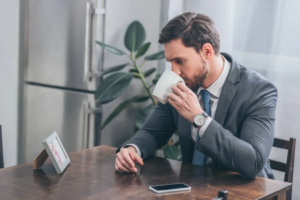 Triste hombre en traje gris sentado en la mesa de madera con el teléfono inteligente beber y mirando poto en el marco en casa, concepto de trastorno de duelo - foto de stock