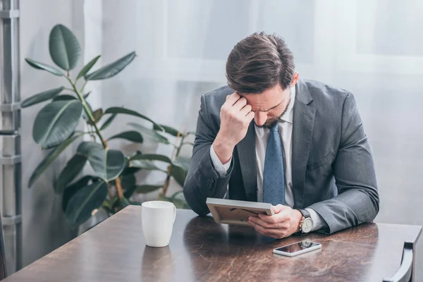 Trauriger Mann im grauen Anzug sitzt am Holztisch mit Smartphone, weißer Tasse und Fotorahmen zu Hause, Trauerbewältigungskonzept — Stockfoto