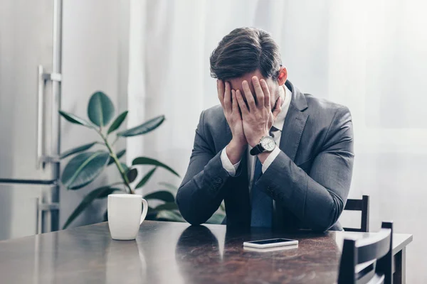 Uomo triste in abito grigio seduto al tavolo di legno con smartphone, cappuccio bianco e faccia di copertura con le mani a casa, concetto di disturbo in lutto — Foto stock