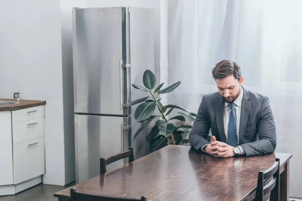 Bouleversé homme en costume gris assis à la table en bois et la pensée dans la cuisine, concept de trouble deuil — Photo de stock