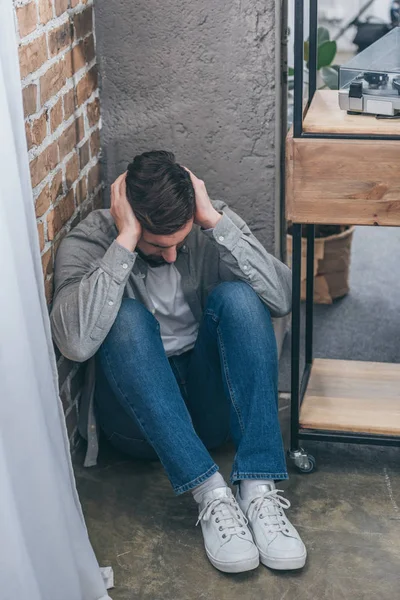 Hombre molesto sentado en el suelo en la esquina y abrazando la cabeza en el fondo de textura marrón en la habitación, concepto de trastorno de duelo - foto de stock