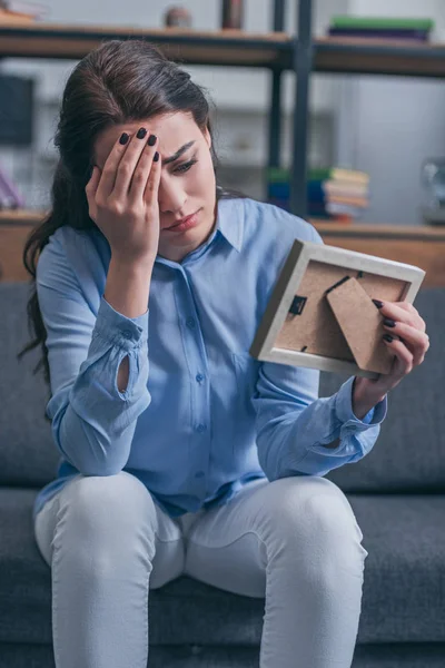 Mulher triste em blusa azul sentado no sofá cinza, olhando para a moldura da foto e chorando em casa, conceito de transtorno de luto — Fotografia de Stock