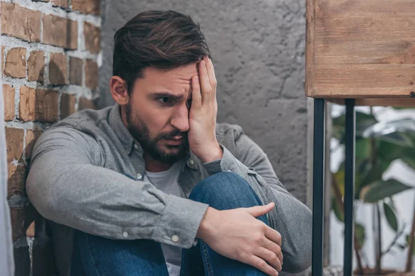 Hombre molesto sentado en la esquina, cubriendo el ojo con la mano sobre el fondo de textura marrón en la habitación, concepto de trastorno de duelo - foto de stock